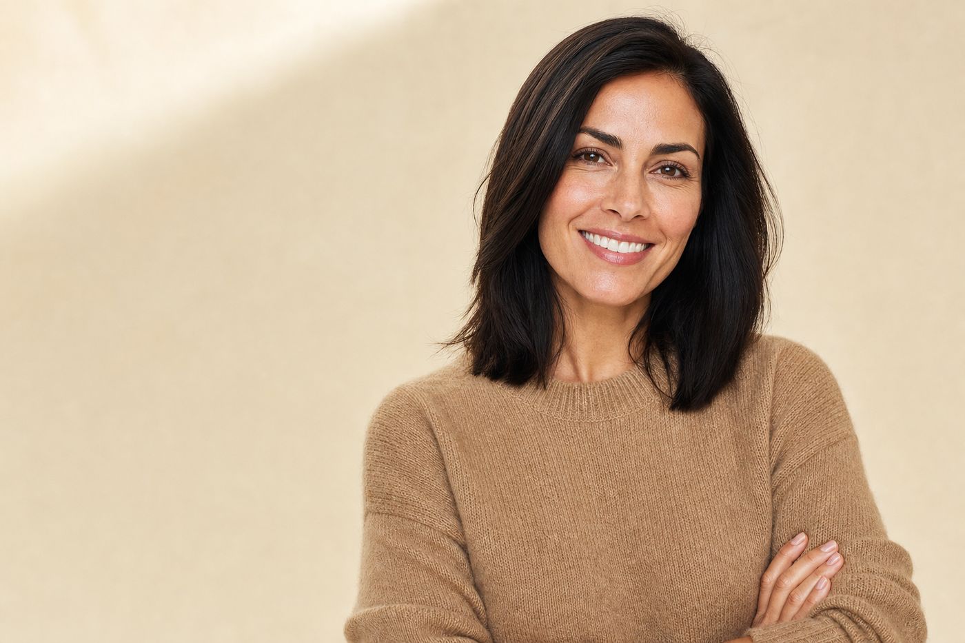 A woman with dark hair in a camel knit, smiling confidently in warm daylight