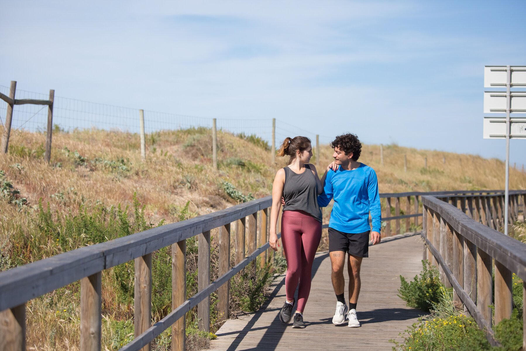 A man and woman walking together outdoors, mid-conversation, on a sunlit boardwalk